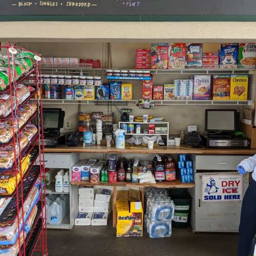 A small convenience store counter with snacks, cereals, and drinks, featuring two cash registers and shelves of products.