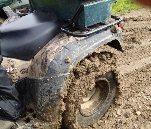 A close-up of a muddy ATV tire, heavily caked with dirt, parked on a rough, unpaved trail.