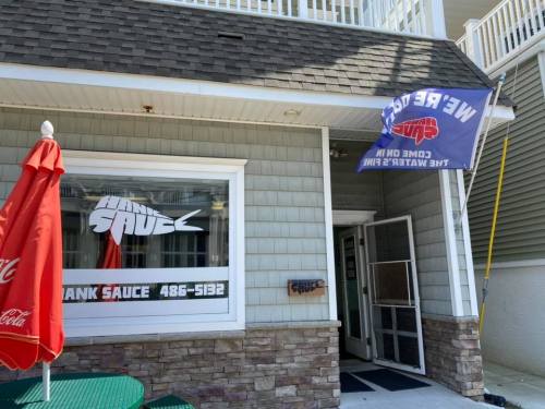 Exterior of a restaurant with a sign reading "Tank Sauce" and a flag that says "We're Open." Red umbrella in front.