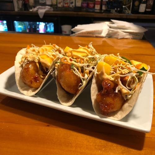 Three tacos filled with crispy fish, topped with cabbage, mango, and a drizzle of sauce, served on a white plate.