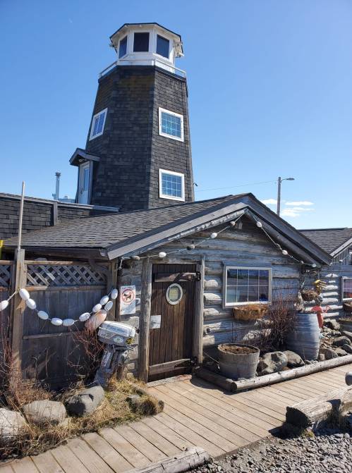 A rustic wooden building with a lighthouse tower, surrounded by rocks and decorative elements, under a clear blue sky.