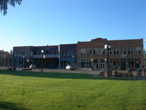 Historic buildings line a street, with a green lawn in the foreground and clear blue skies above.