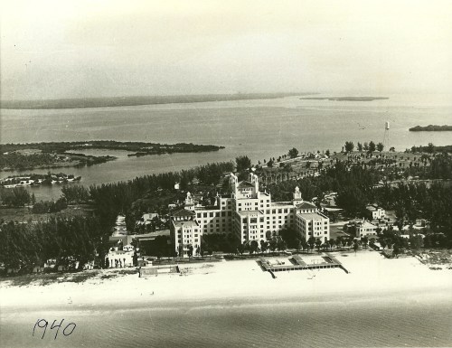 Aerial view of a large hotel by the beach in 1940, surrounded by water and lush greenery.