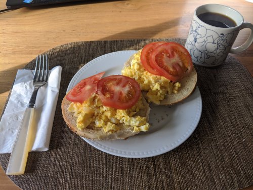 A plate with scrambled eggs on a toasted bun, topped with tomato slices, alongside a cup of coffee.