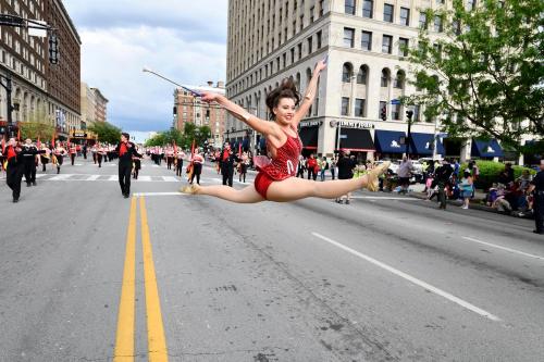 A performer in a red costume leaps gracefully in a parade, with marching bands and spectators in the background.