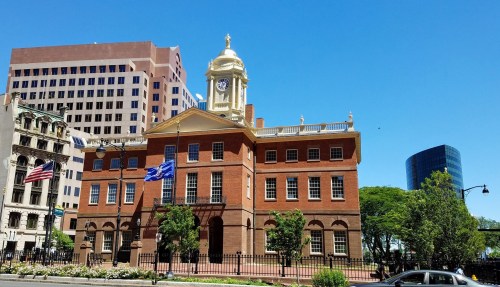 Historic building with a clock tower, surrounded by greenery and flags, under a clear blue sky.