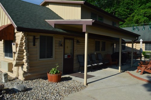 A cozy log cabin-style house with a porch, surrounded by greenery and gravel landscaping.