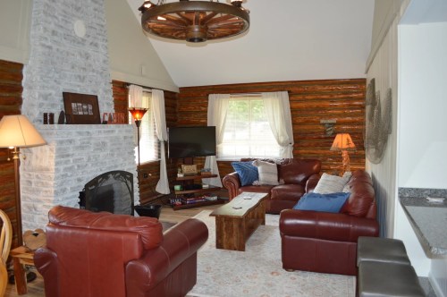 Cozy living room with leather sofas, a stone fireplace, and rustic decor, featuring a TV and large windows.
