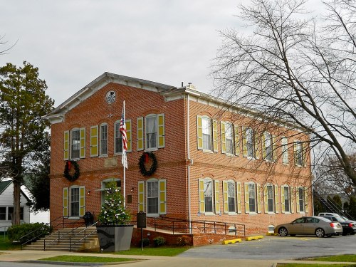 A brick building with yellow shutters, holiday wreaths, and a small Christmas tree outside, set against a cloudy sky.