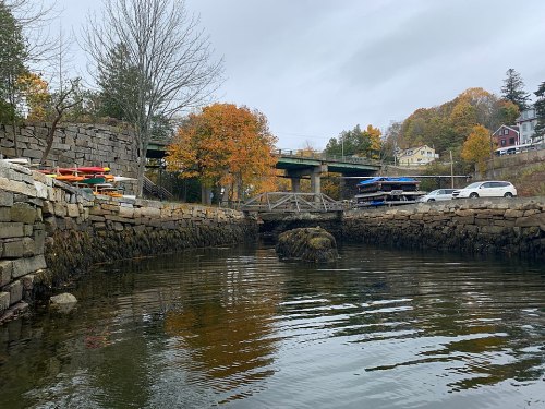 A calm waterway with a stone bridge, autumn trees, and colorful kayaks under a cloudy sky.
