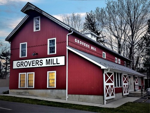 A red barn-style building with "GROVERS MILL" signage, surrounded by trees and a clear sky.