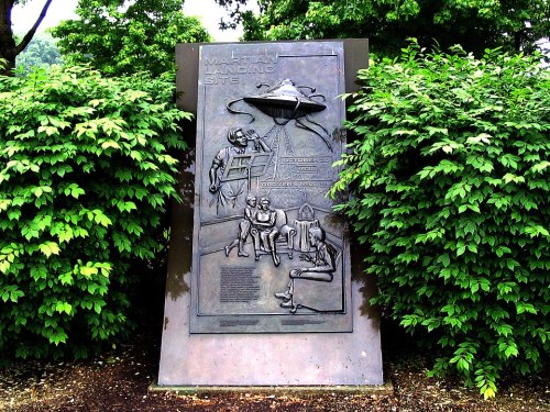 A bronze plaque depicting a UFO landing scene, surrounded by lush green foliage.
