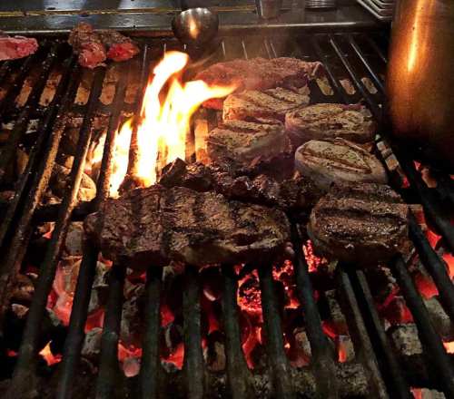 Grilling various cuts of meat over an open flame, with vibrant flames and glowing coals visible.
