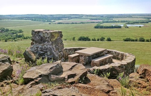 A stone picnic area on a hilltop, overlooking a lush green landscape and distant water bodies under a clear blue sky.