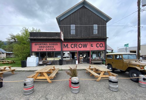 A rustic storefront with "M. Crow & Co." sign, featuring picnic tables and an old vehicle parked outside.