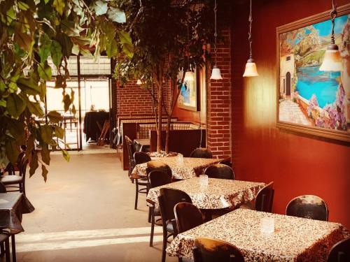 Cozy restaurant interior with tables covered in patterned cloth, greenery, and warm lighting. Brick walls and artwork visible.
