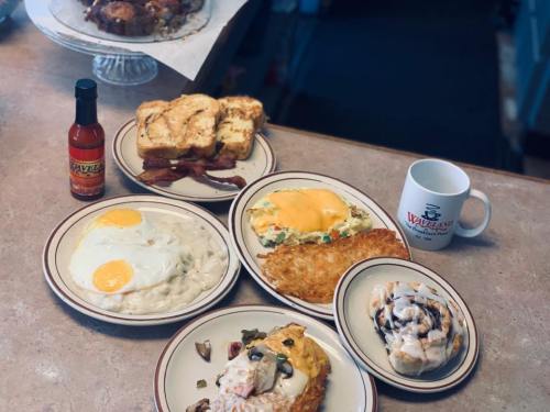 A spread of breakfast dishes including eggs, bacon, hash browns, toast, and a cinnamon roll, with a coffee mug and hot sauce.