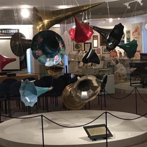 Colorful vintage gramophone horns suspended in a museum display, surrounded by various artifacts and framed pictures.