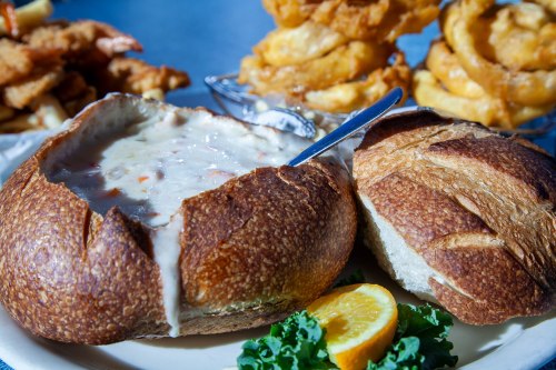 A bread bowl filled with creamy soup, garnished with a lemon slice and served with fried shrimp and onion rings in the background.