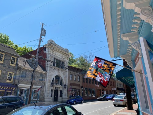 A sunny street scene featuring a Maryland flag, historic buildings, and parked cars under a clear blue sky.