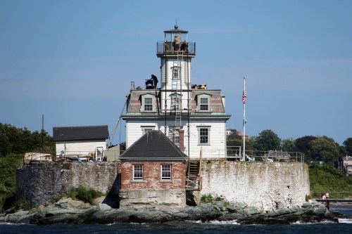 A historic lighthouse on a rocky shore, with workers on scaffolding and a clear blue sky in the background.