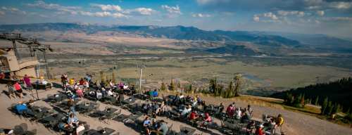 A scenic mountain view with people dining at outdoor tables, overlooking valleys and distant mountains under a blue sky.
