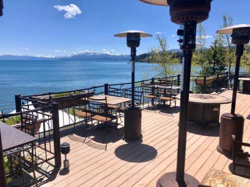 Scenic lakeside patio with tables, heaters, and mountains in the background under a clear blue sky.