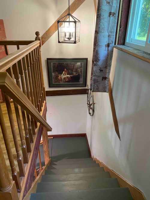 A staircase leading down, with a lantern hanging above and a framed picture on the wall. Natural light from a window.