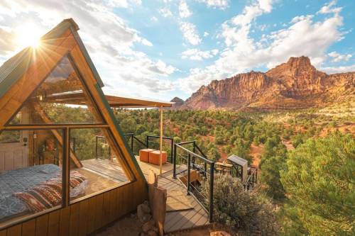 A cozy A-frame cabin with large windows overlooks a scenic desert landscape and mountains under a bright sky.