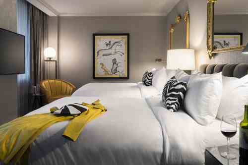 A stylish hotel room with two beds, a modern chair, artwork, and a glass of wine on a bedside table.