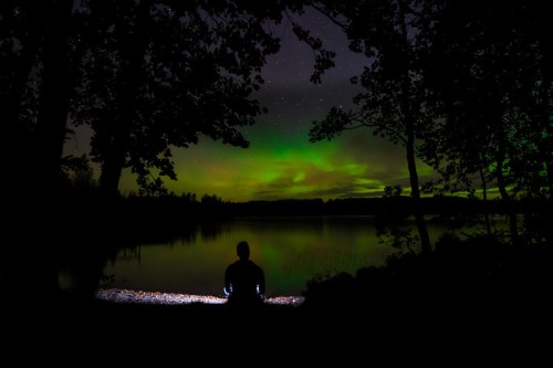 A person sits by a calm lake at night, illuminated by a soft light, with vibrant green auroras in the sky above.