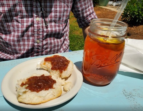 A plate with two biscuits topped with jam, next to a jar of iced tea on a sunny outdoor table.