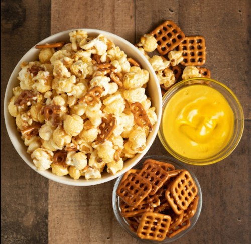 A bowl of popcorn mixed with pretzels, accompanied by a small dish of yellow cheese dip and a jar of pretzels.
