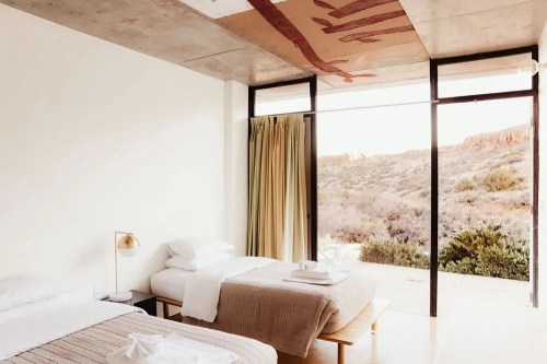 A modern bedroom with two beds, large windows, and a view of a natural landscape outside.