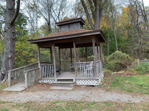 A wooden gazebo surrounded by trees, featuring a sloped roof and open sides, with a path leading to it.