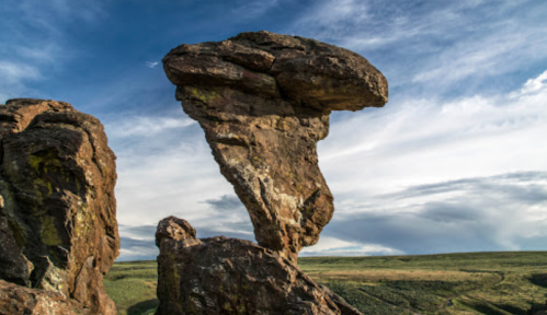 A tall, thin rock formation stands against a blue sky with wispy clouds, surrounded by green hills.