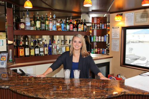 A smiling bartender stands behind a bar with various bottles of liquor and drinks displayed on shelves.