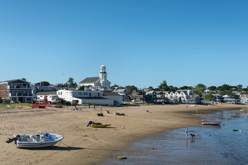 A sunny beach scene with boats on the sand, a church tower, and coastal buildings in the background.