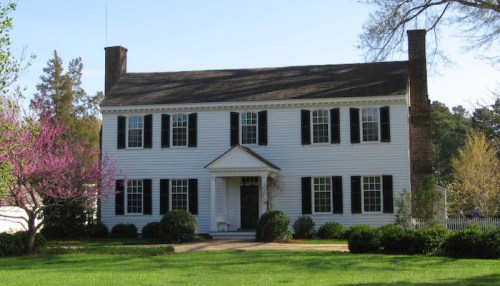 A classic two-story white house with black shutters, surrounded by green grass and blooming trees.