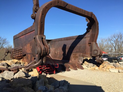 A large, rusted excavator bucket displayed outdoors, surrounded by rocks and a clear blue sky.