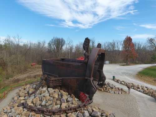 A large, rusted metal sculpture resembling a mining tool, surrounded by rocks and trees under a blue sky.