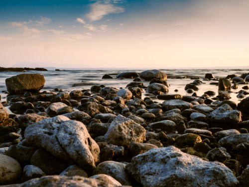 A rocky shoreline at sunset, with smooth stones and gentle waves under a colorful sky.
