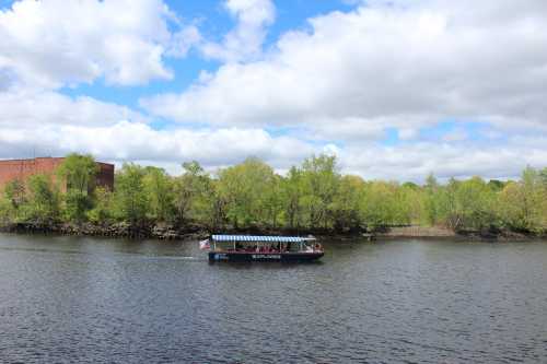 A tour boat glides along a river, surrounded by lush green trees and a partly cloudy sky.