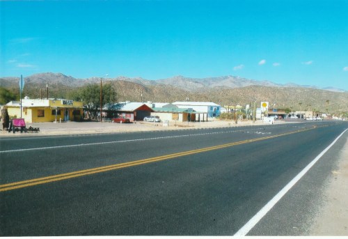 A wide view of a desert road lined with small buildings and mountains in the background under a clear blue sky.
