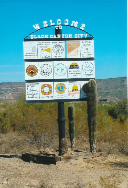 Welcome sign for Black Canyon City, featuring various local logos and a backdrop of desert vegetation.
