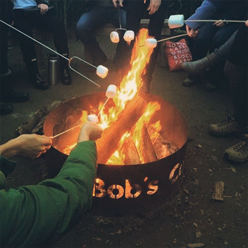 A group of people roasting marshmallows over a campfire in a metal fire pit labeled "Bob's."