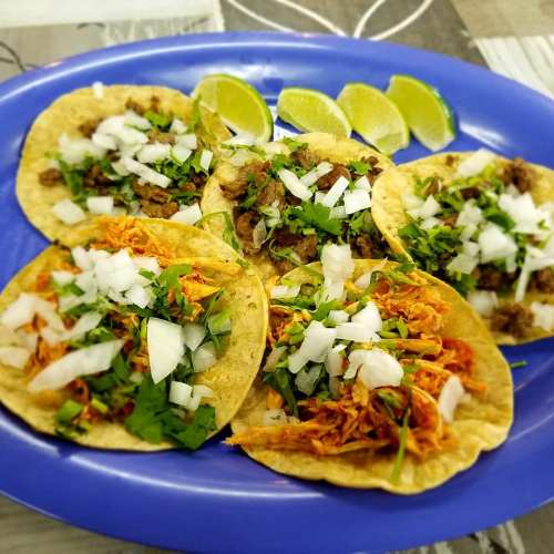 A blue plate with six tacos topped with onions and cilantro, accompanied by lime wedges.
