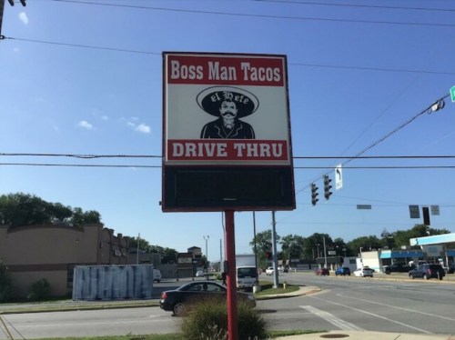 Sign for "Boss Man Tacos" with a drive-thru, featuring a cartoonish taco character and a clear blue sky.