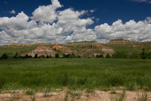 Vast green field in the foreground with colorful hills and a dramatic sky filled with clouds in the background.