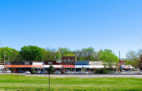 A small town street with shops, trees, and a clear blue sky in the background. Green grass in the foreground.